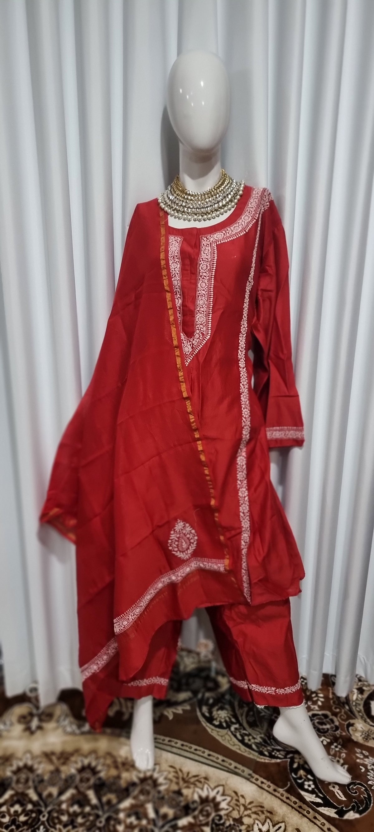 A red traditional South Asian outfit displayed on a white mannequin, featuring a kurta with white embroidery, matching pants, and a draped dupatta, set against a white curtain background.